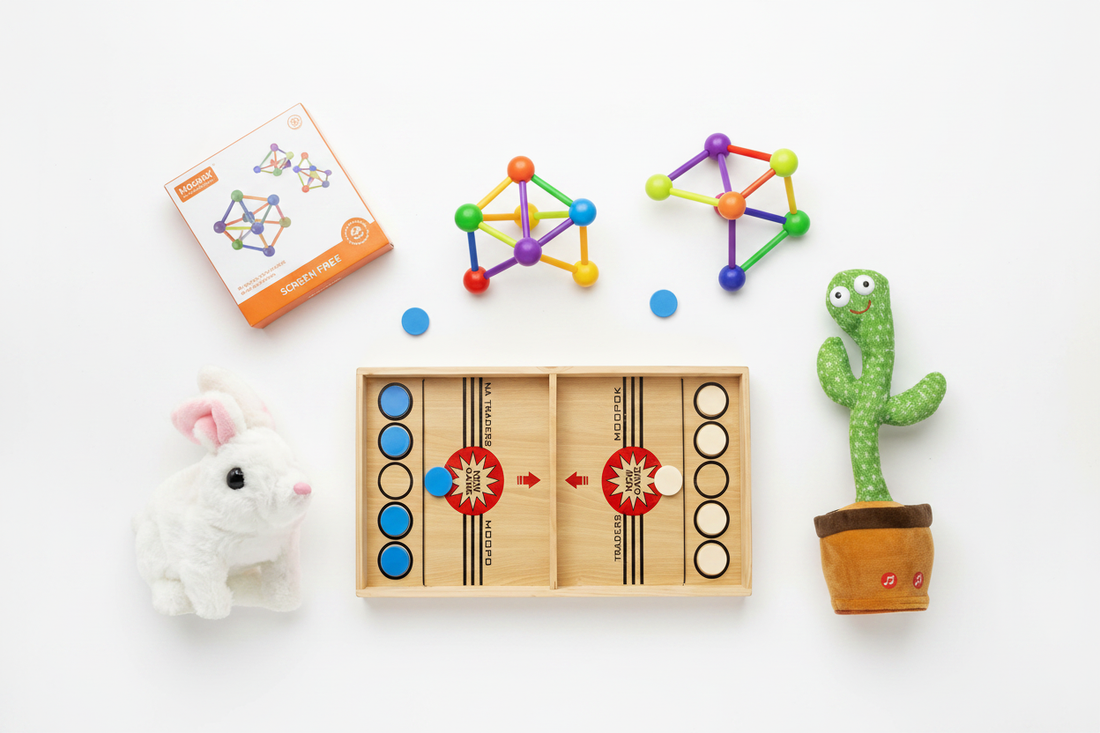 Flat lay of Go Baby screen-free kids toys including magnetic tiles, wooden sling puck game, plush rabbit, and dancing cactus on white background